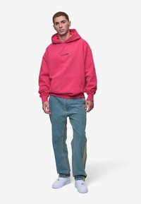 Rozā kapuce ar "PEGADOR" logotipu, kengura kabata, ribotās aproces, saskaņotas ar gaiši ziliem džinsiem un baltām sporta apaviem. Minimālistisks dizains.