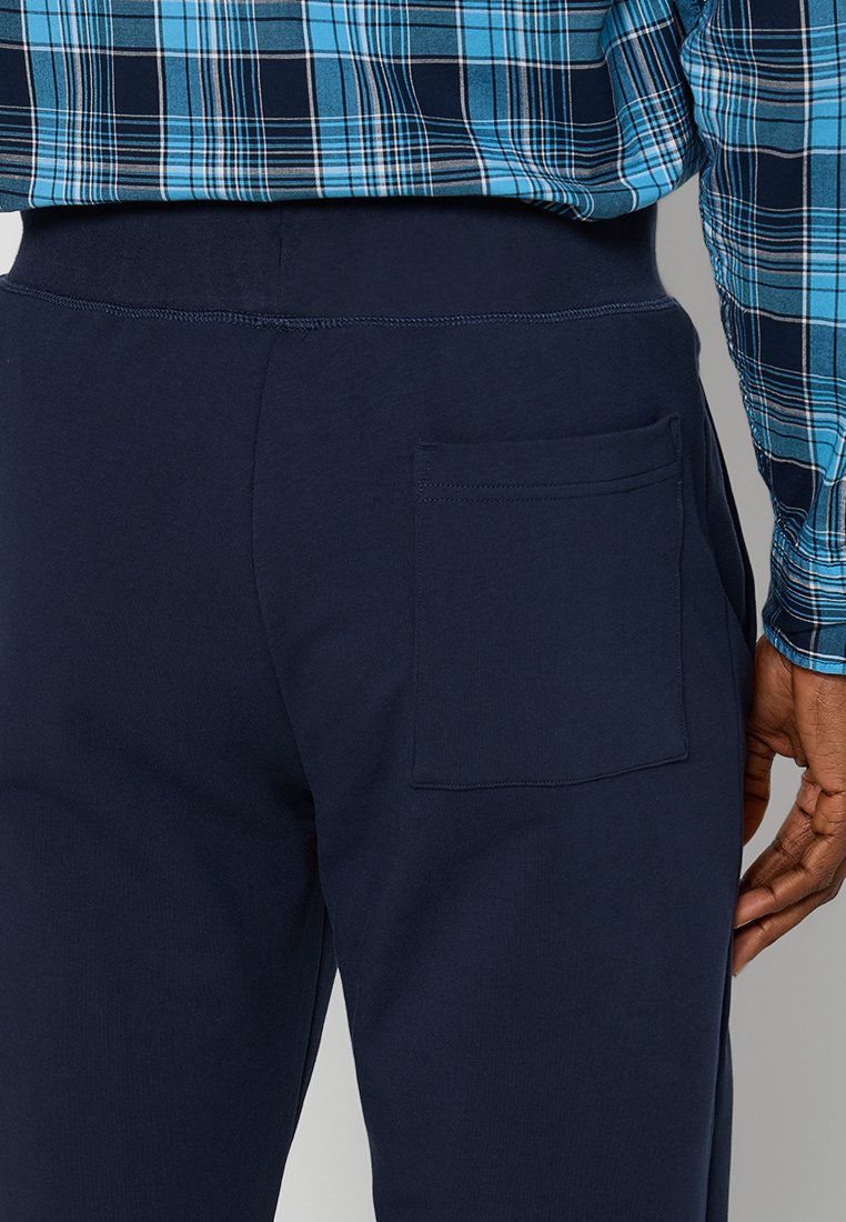 Uomo con pantaloni blu navy dotati di una tasca posteriore e una camicia a quadri blu infilata dentro, con una mano appoggiata di lato.