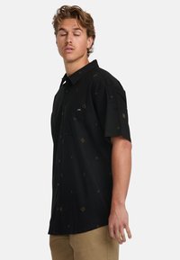 Camisa negra de manga corta con botones, que presenta patrones geométricos dorados, un bolsillo en el pecho y un corte relajado con una textura de tela suave.