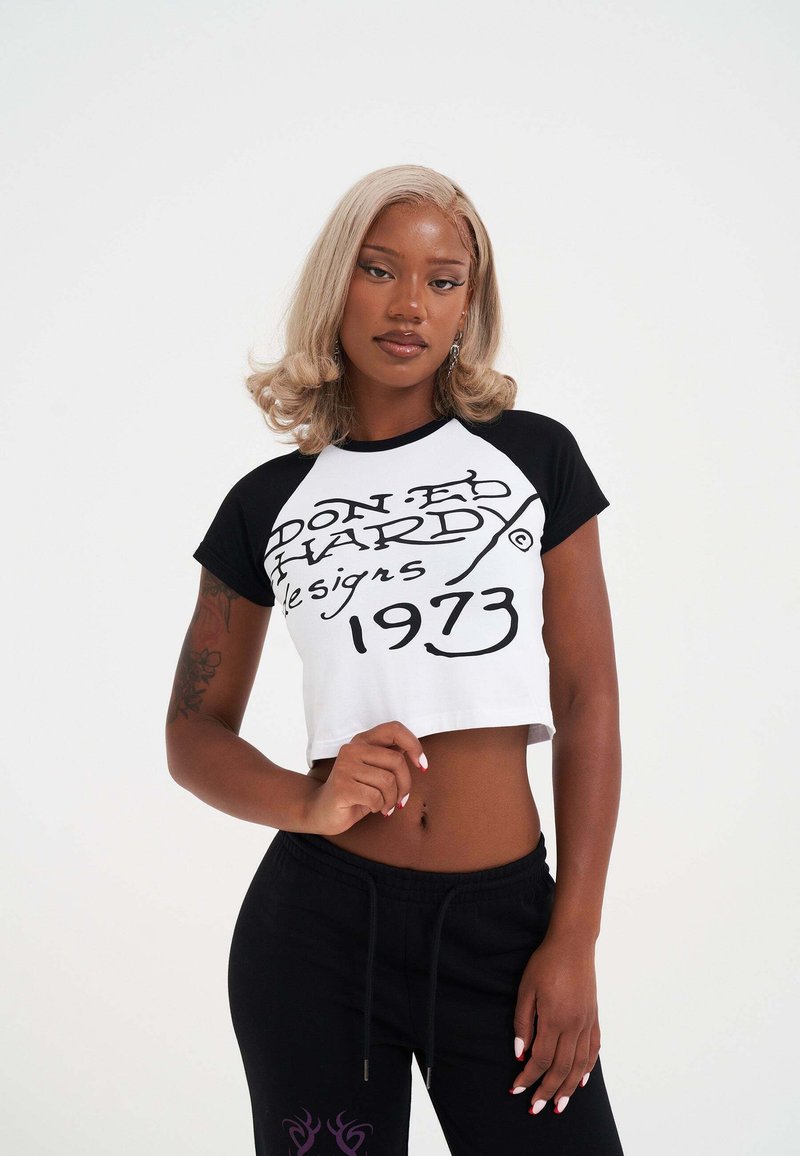 Kobieta z blond kręconymi włosami, ubrana w czarno-białą, krótką koszulkę Don Ed Hardy z 1973 roku i czarne spodnie, stojąca na tle czystego białego tła.