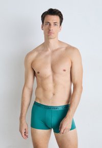 Moške boksarice Calvin Klein v živahni teal barvi z mehkobno teksturo, ki imajo črn logotip na pasu in prilegajoč dizajn.