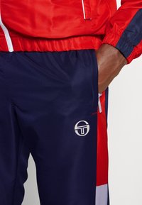Chaqueta roja con cremalleras, combinada con pantalones azul marino que presentan un logo blanco. Tejido texturizado, puños y cintura elásticos, y detalles de bloqueo de color.
