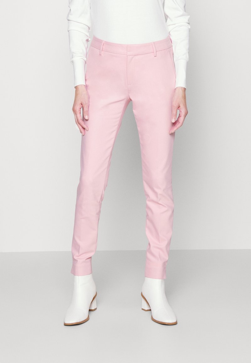 Femme portant un pantalon slim rose clair et des bottines blanches, debout devant un fond clair uni.