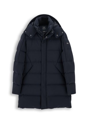 Homme portant un bonnet gris, un long manteau matelassé foncé, une chemise bleue, un cardigan gris, un jean et des baskets bleues, debout les mains dans les poches du manteau.