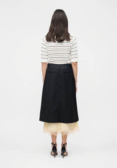 Γυναίκα που στέκεται με την πλάτη, φορώντας λευκό ριγέ τοπ, μαύρη μίντι φούστα επάνω από μπεζ δαντελένιο φόρεμα και μαύρα σανδάλια με τακούνια.