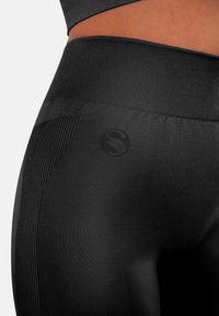 Leggings neri a vita alta con una texture liscia e elastica, caratterizzati da sottili dettagli a coste e un logo stampato sul lato inferiore.