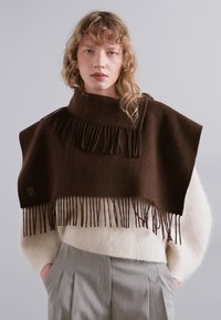 Poncho brun à franges en laine douce, avec un col montant. Associé à un pull crème et un pantalon plissé gris clair.