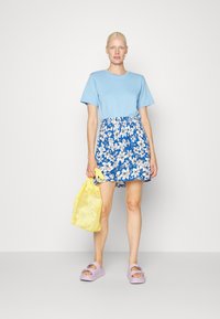 T-shirt de algodão azul combinado com uma saia azul estampada com flores, uma mala de rede amarela e sandálias de plataforma lavanda.
