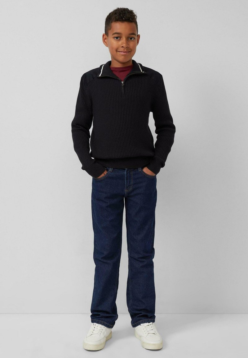 Schwarzer Strickpullover mit Reißverschlusskragen, kombiniert mit blauen Denim-Jeans und weißen Sneakers. Verfügt über eine gerippte Textur und kontrastierende Kragendetails.