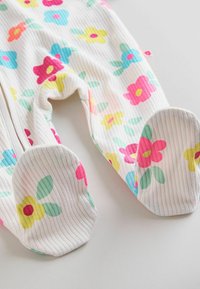 Pyjama bébé blanc côtelé à pieds avec un imprimé de fleurs colorées en rose, jaune et bleu, étalé à plat sur une surface claire.