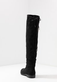 Stivale sopra al ginocchio in suede nero con una texture liscia, punta arrotondata e suola piatta. Caratterizzato da un dettaglio con lacci nella parte superiore e una zip laterale.