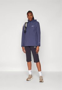 Navy waterproof jacket with a hood, paired with black knee-length shorts. Model wears beige sneakers and white ankle socks.