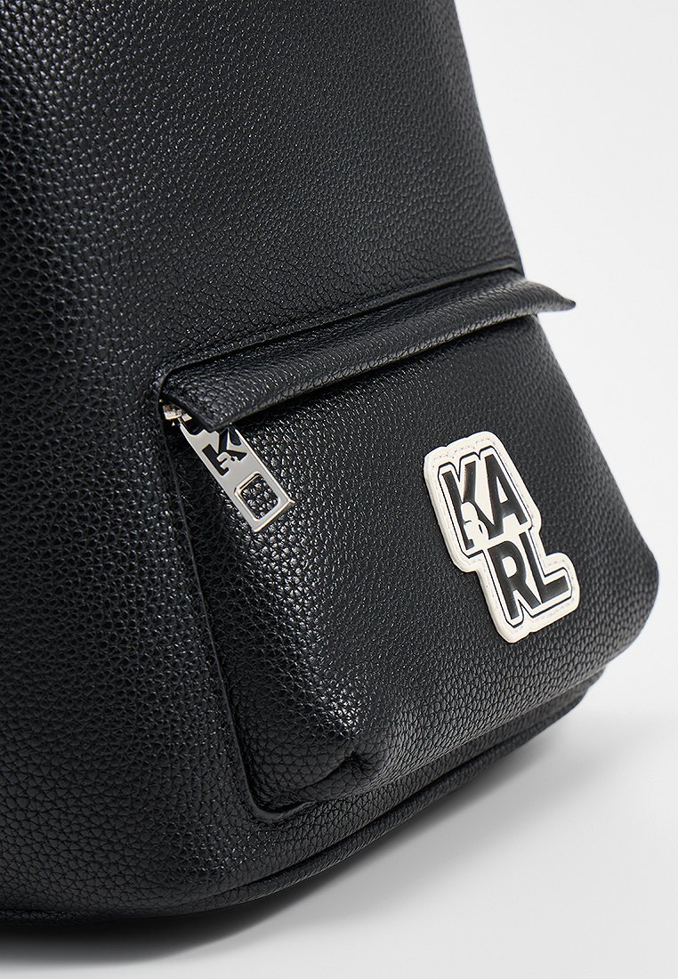 Czarna skórzana torba o teksturowanej powierzchni, z kieszenią i srebrnym zamkiem błyskawicznym oraz białą łatą z logo "KARL" w wyraźnych czarnych literach.