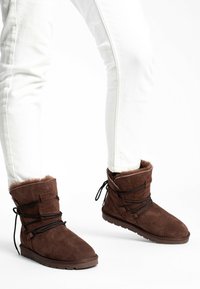 Stivaletti in suede marrone con fodera in pelliccia sintetica, caratterizzati da lacci incrociati e suola piatta in gomma. Indossati con jeans bianchi attillati.
