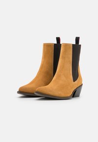 Tan mockasinstövlar med svarta elastiska sidopaneler och en kort blockklack. Har en dragflik bak och en spetsig tådesign.