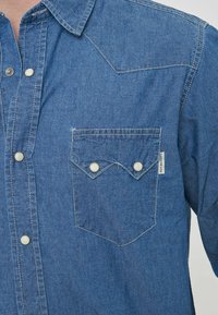 Camicia di denim con maniche lunghe, colore azzurro chiaro, due bottoni sul petto e una tasca con bavetta appuntita. Presenta cuciture e etichette visibili.