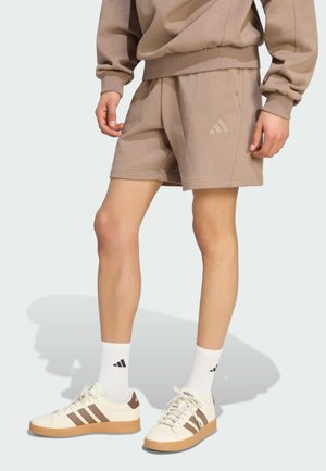 Personne portant un short beige Adidas, un sweat assorti, des chaussettes blanches Adidas et des baskets blanches avec des bandes marron et des semelles en gomme.
