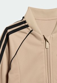 Bež suknjič z zadrgo, črne črte na ramenih, rebrast ovratnik in gladka tekstura tkanine. Na prsih ima majhen logotip.