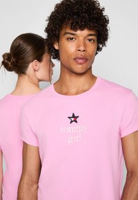 Rožinė medvilninė marškinėliai su apvaliu kaklu, trumpomis rankovėmis ir siuvinėta juoda žvaigžde bei "tommy girl" tekstu priekyje.