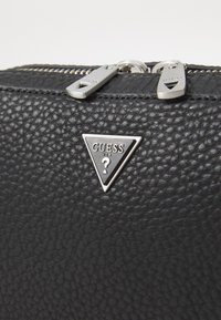 Schwarze geprägte Ledertasche mit einem silbernen dreieckigen Guess-Logo und zwei Metallreißverschlüssen. Die ausgeprägte strukturierte Oberfläche ist markant.
