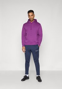 Lilla hættetrøje med justerbar snor og frontlomme, parret med marineblå sweatpants med et hvidt logo, og sorte træningssko.