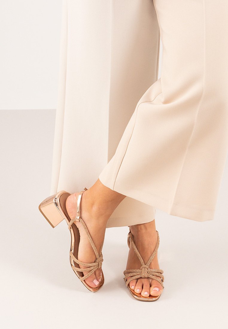 Sandalias beige de tiras con tacón cuadrado, que presentan un diseño trenzado y acentos metálicos brillantes en el tacón y las tiras.