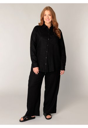 Vrouw met lang rood haar lacht, draagt een losse zwarte blouse met knopen, brede zwarte broek en zwarte slidesandalen, staand tegen een effen achtergrond.