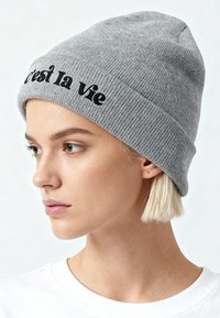 Jeune femme aux cheveux blonds courts portant un bonnet gris en maille brodé de "c'est la vie" en noir, faisant face à gauche, vêtue d'une chemise blanche.
