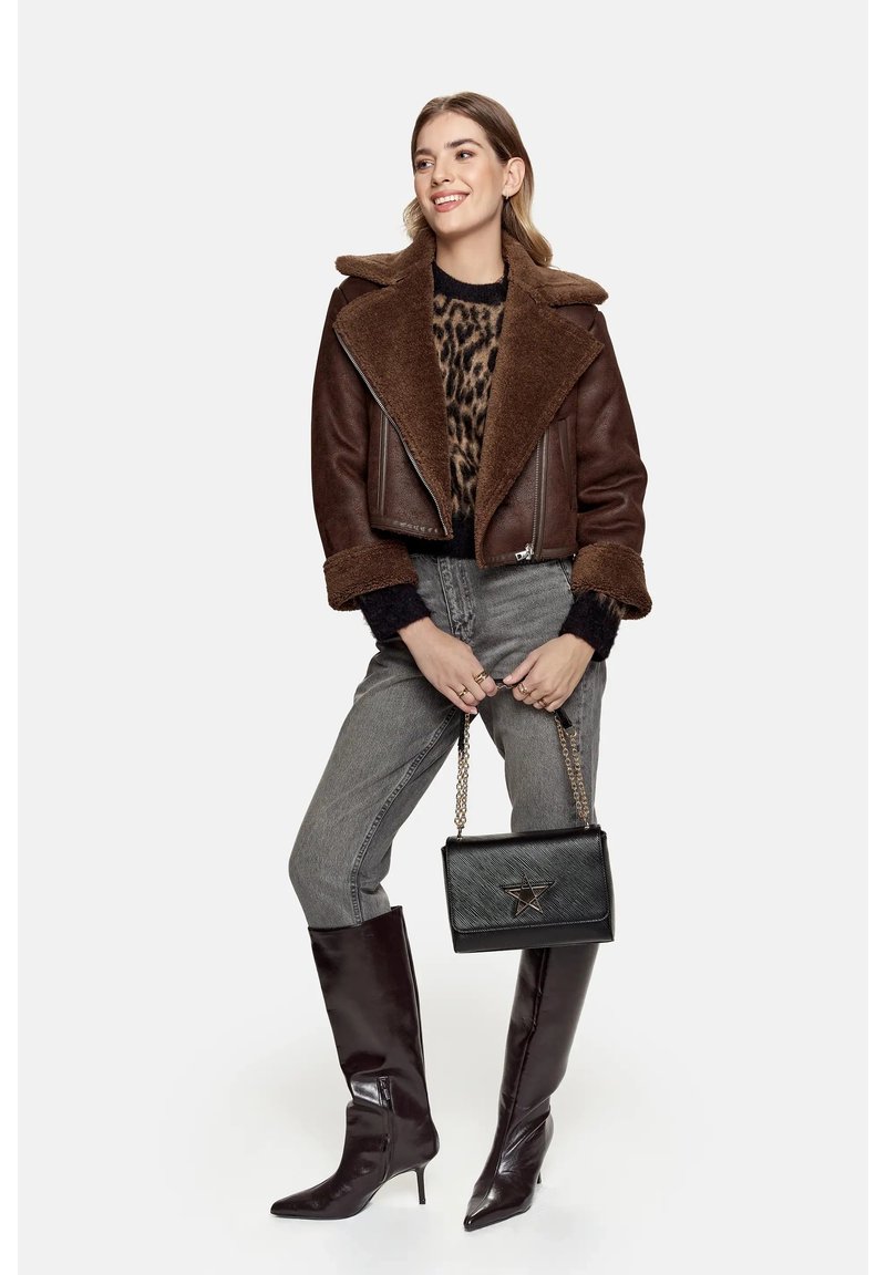 Brun kort shearlingjacka med bred krage, leopardmönstrad tröja, grå jeans, glänsande burgundy knähöga stövlar och en svart handväska med stjärndetalj.