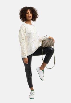 Kvinde med krøllet hår iført hvid kabelstrikket sweater og sorte læderbukser, der holder en quiltet olivengrøn crossbody-taske.