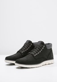 Svart mockastonknäboots med grå detaljer, som har en vadderad krage, metallöglor och en vit sula. Logotypen präglad på sidan.
