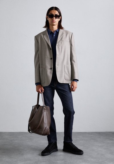 Ljusgrå blazer med två fickor, marinblå polo-t-shirt, mörka jeans, svarta skor och en brun läderväska med silverdetaljer.