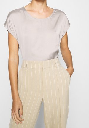 Vrouw draagt een lichtgrijze blouse met korte mouwen en ronde hals en beige broek met hoge taille en witte verticale strepen, linkerhand in de broekzak.