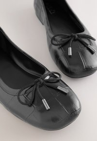 Ballerines en cuir verni noir avec bouts carrés et nœuds décoratifs en cordon noir avec extrémités argentées sur une surface claire.