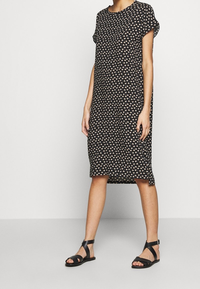 Femme portant une robe noire longueur genou avec de petits motifs triangulaires blancs et beiges et des sandales plates noires à brides, debout devant un fond uni.