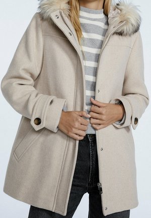 Manteau en laine beige avec capuche bordée de fourrure, poignets boutonnés, fermetures éclair et pressions, porté sur un pull rayé gris et blanc.