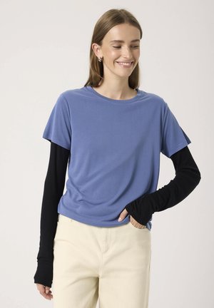 T-shirt s krátkým rukávem modré barvy vrstvený přes dlouhé černé rukávy. Měkká látka, kulatý výstřih, volný střih, nošeno s kalhotami světlé barvy.