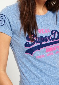 Ljusblå kortärmad t-shirt med rund halsringning, med mörkblå och rosa grafisk text samt siffran "8" på vänster ärm.