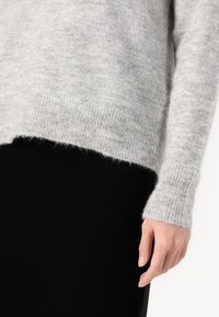 Main portant une manche de pull en tricot duveteux gris clair sur un pantalon noir, visible de la taille à la main.