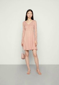 Mulher vestindo um vestido longo de renda rosa, segurando uma mala rosa combinando, em pé sobre um chão cinza contra uma parede clara.