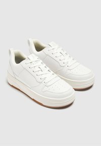 Un paio di sneakers bianche in pelle con chiusura a lacci, punta perforata, colletto imbottito e suole in gomma marrone su uno sfondo grigio chiaro.