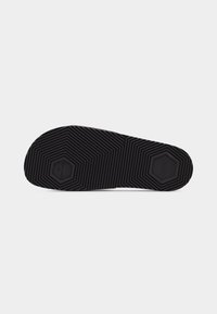 Semelle de chaussure en caoutchouc noir avec des motifs hexagonaux et des rainures texturées pour une meilleure adhérence, présentée sur un fond gris clair uni.