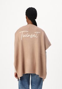 Poncho lavorato a maglia beige chiaro con vestibilità ampia, caratterizzato da un grande logo bianco "Twinset" ricamato sul retro. Tessuto morbido e lati aperti.