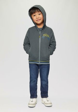Graue Zip-Hoodie mit Kapuze, mit mehrfarbigem Textdesign, kombiniert mit blauen Jeans und weißen Sneakers mit grauen Akzenten.