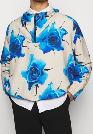 Beige pullover med blå rosenmønstre, båret over en hvid skjorte, kombineret med sorte bukser, hænderne afslappet i lommerne.