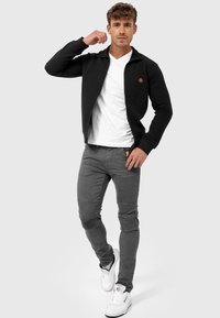 Sort textured zip-up jakke med et rundt logo, parret med grå slim-fit jeans med ribbede detaljer. Hvide sneakers fuldender looket.