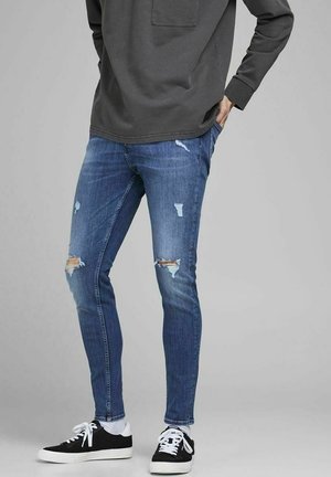 Jean denim bleu avec des détails usés, présentant des déchirures et des décolorations. Associés à des baskets noires et des chaussettes blanches pour le contraste.