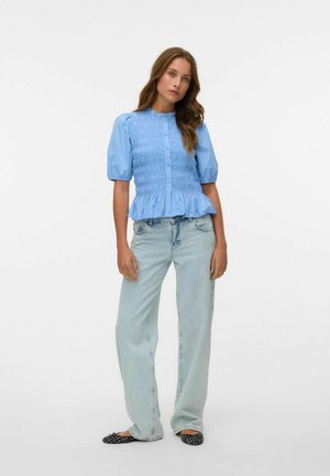 Blouse bleu clair à manches bouffantes avec un devant smocké et un ourlet volanté, associée à un jean large bleu clair et des chaussures plates noires.