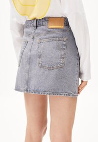 Jupe en denim bleu clair, à ourlet brut, avec deux poches arrière, coutures visibles et une étiquette en cuir marron portant l'inscription "ARMEDANGELS."