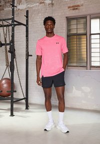 Camisola desportiva rosa de manga curta, com logótipo no peito, combinada com calções pretos. Modelo em pé num ginásio com equipamento de fitness.